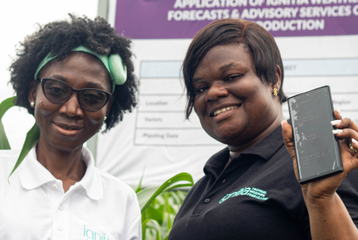 two women farmers standing in crop field, smiling and holding up mobile phones